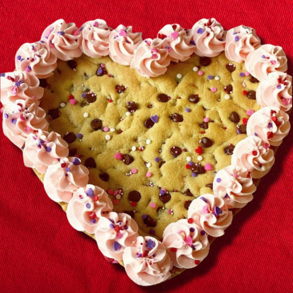 Heart-shaped cookie cake with pink frosting and colorful sprinkles on a red background.