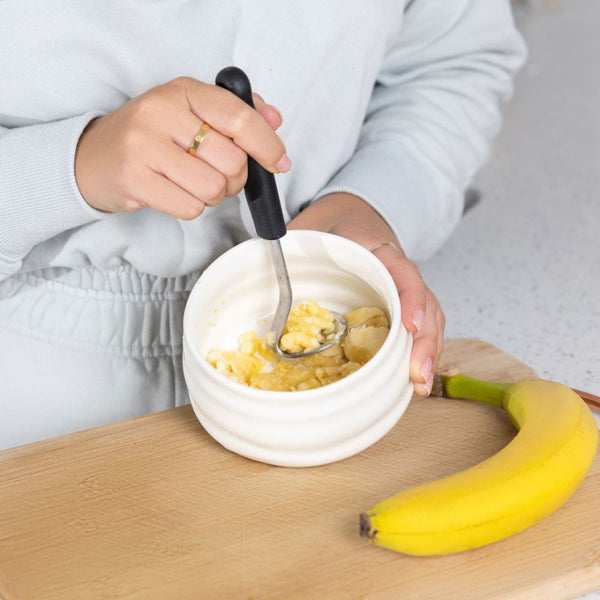 Person using a mini masher with black handle to mash bananas in a white bowl on a wooden cutting board.